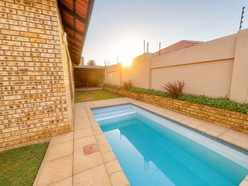 A well-maintained outdoor pool area featuring a paved surround, brick boundary walls with electric fencing, and a covered built-in braai area.