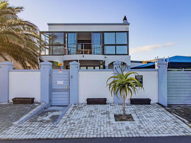 Front elevation of a modern two-story house featuring a secure perimeter wall, gated entrance, paved driveway, and a balcony with glass balustrades.