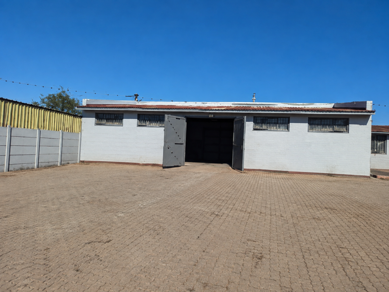 Exterior view of a commercial warehouse building featuring a large paved yard, secure metal entrance gates, and security-barred windows.