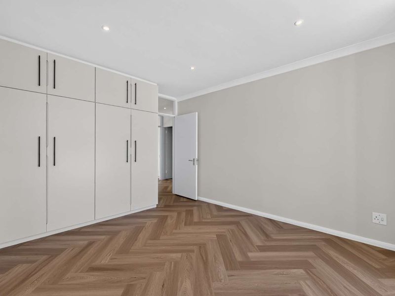 A bright, empty bedroom featuring modern herringbone flooring, neutral walls, and extensive built-in cabinetry with recessed lighting.