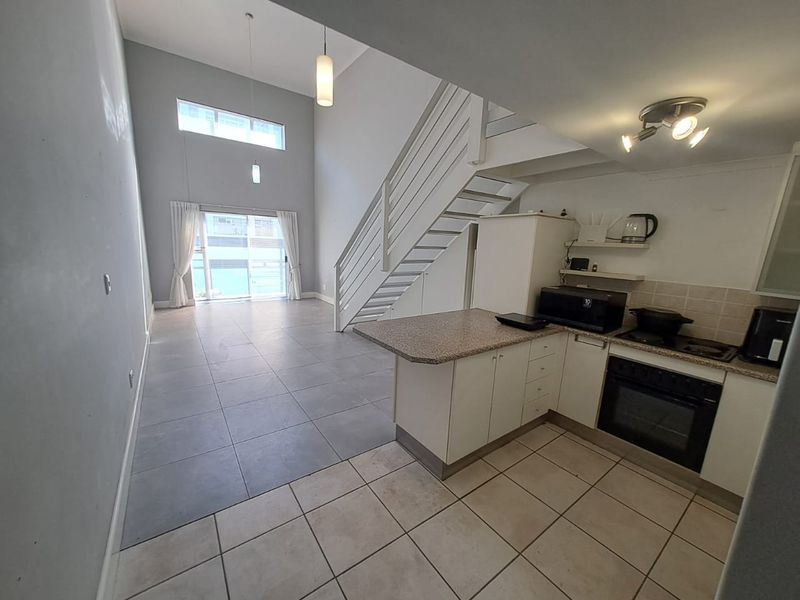 Open-plan living area featuring a modern staircase, tiled flooring, and a functional kitchen with built-in oven and hob, leading to a bright balcony.