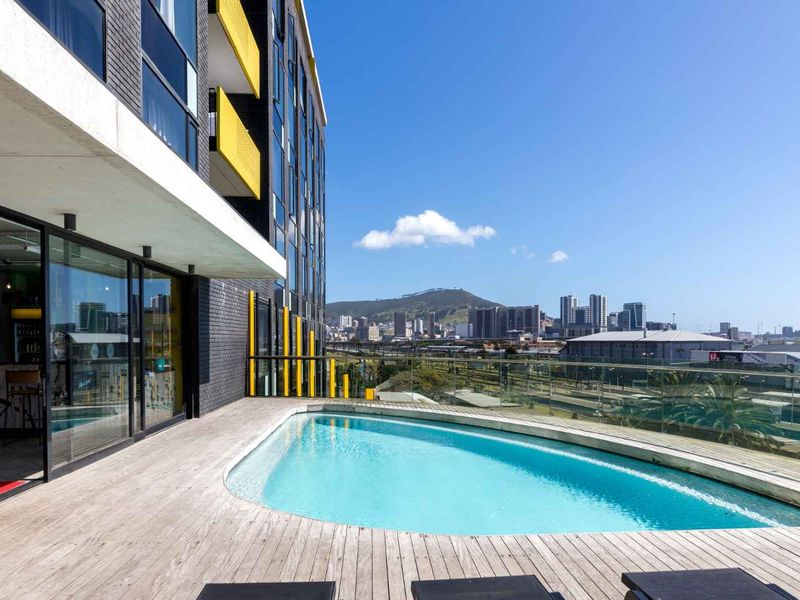 A modern rooftop pool area featuring timber decking, glass balustrades, and expansive city and mountain views.