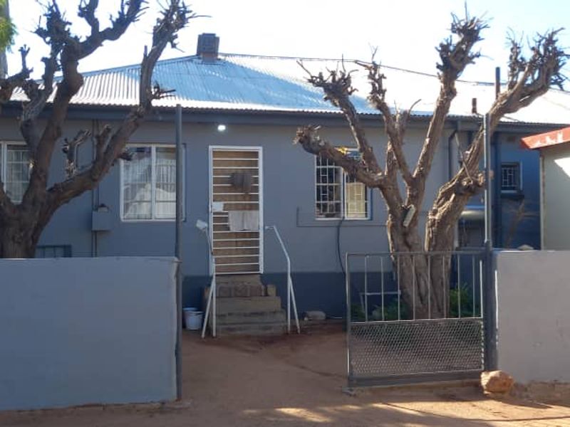 Front elevation of a single-story house featuring a corrugated metal roof, security gates, and a perimeter wall with a metal entrance gate.
