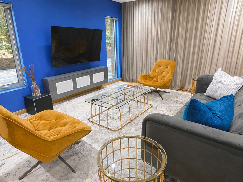 A modern living room featuring a bold blue accent wall, large windows with floor-to-ceiling curtains, and a wall-mounted television setup.