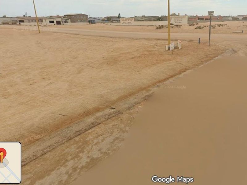A street-level view of a vacant residential plot in a sandy, undeveloped coastal area with existing street lighting infrastructure.