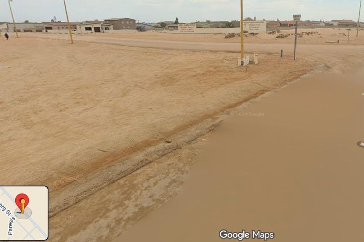 A street-level view of a vacant residential plot in a sandy, undeveloped coastal area with existing street lighting infrastructure.