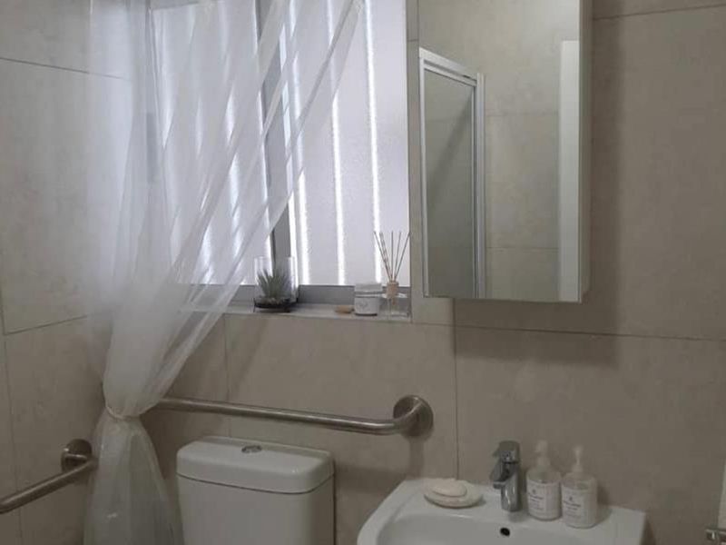 A functional bathroom featuring a wall-mounted basin, toilet, mirrored medicine cabinet, and stainless steel grab rails, finished with neutral wall tiles.