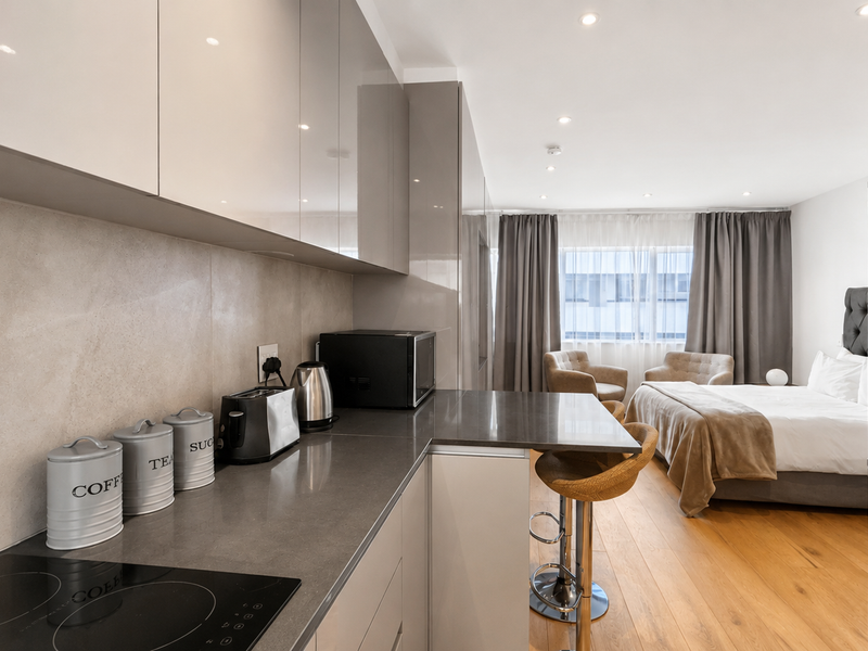 Modern open-plan studio apartment featuring a sleek kitchen with glossy cabinetry, stone countertops, and an integrated fridge, flowing into a comfortable bedroom area.