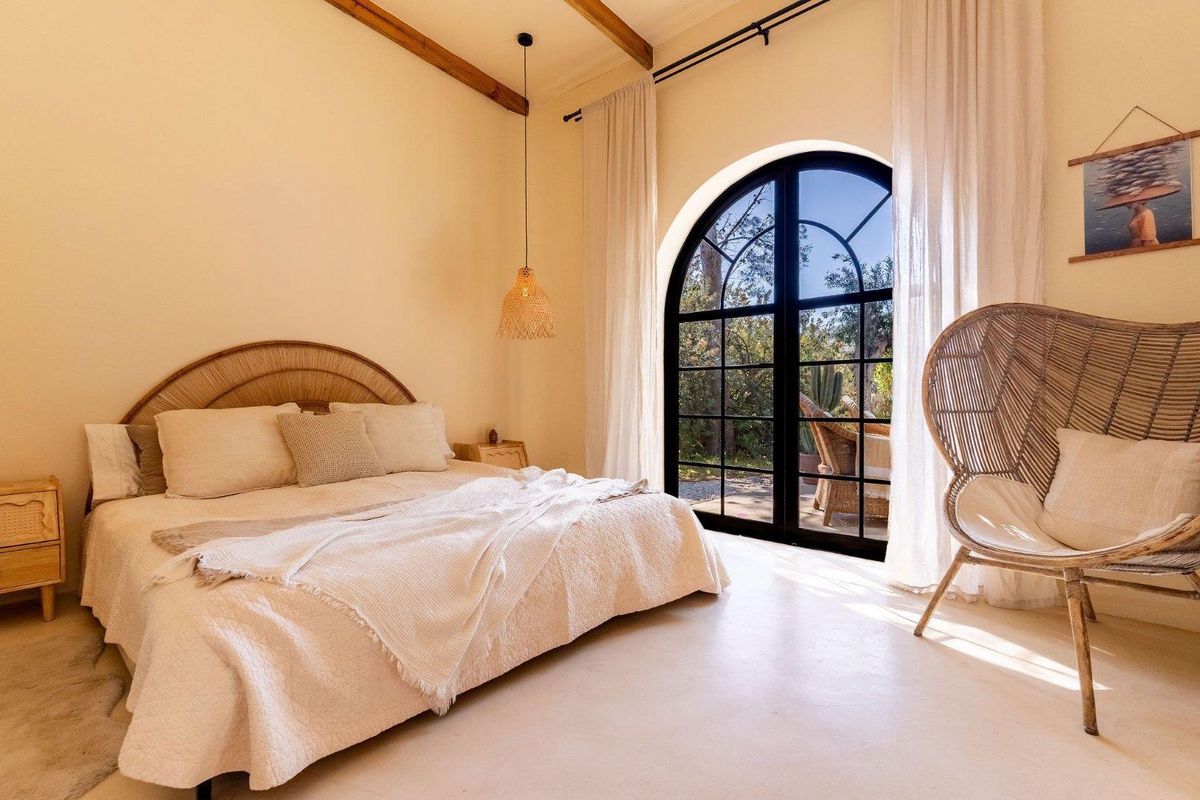 A bright bedroom featuring exposed timber ceiling beams, polished concrete flooring, and a large arched black-framed glass door providing direct access to the garden.