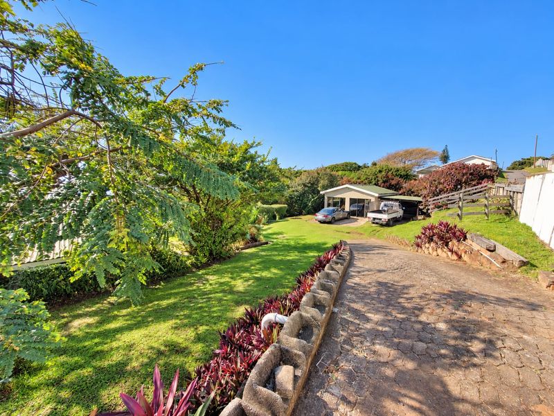 A wide-angle view of the property entrance featuring a paved driveway, landscaped garden beds, and a carport structure.
