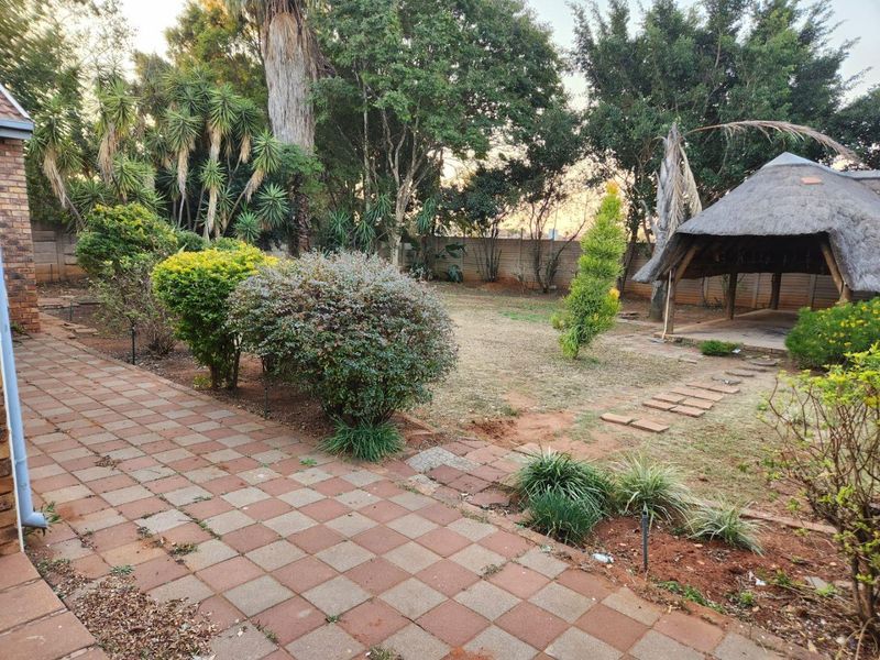 A spacious rear garden featuring a paved patio area, established trees, and a traditional thatched lapa structure, offering potential for outdoor landscaping.