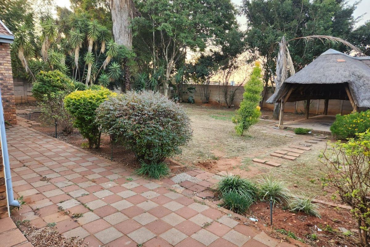 A spacious rear garden featuring a paved patio area, established trees, and a traditional thatched lapa structure, offering potential for outdoor landscaping.