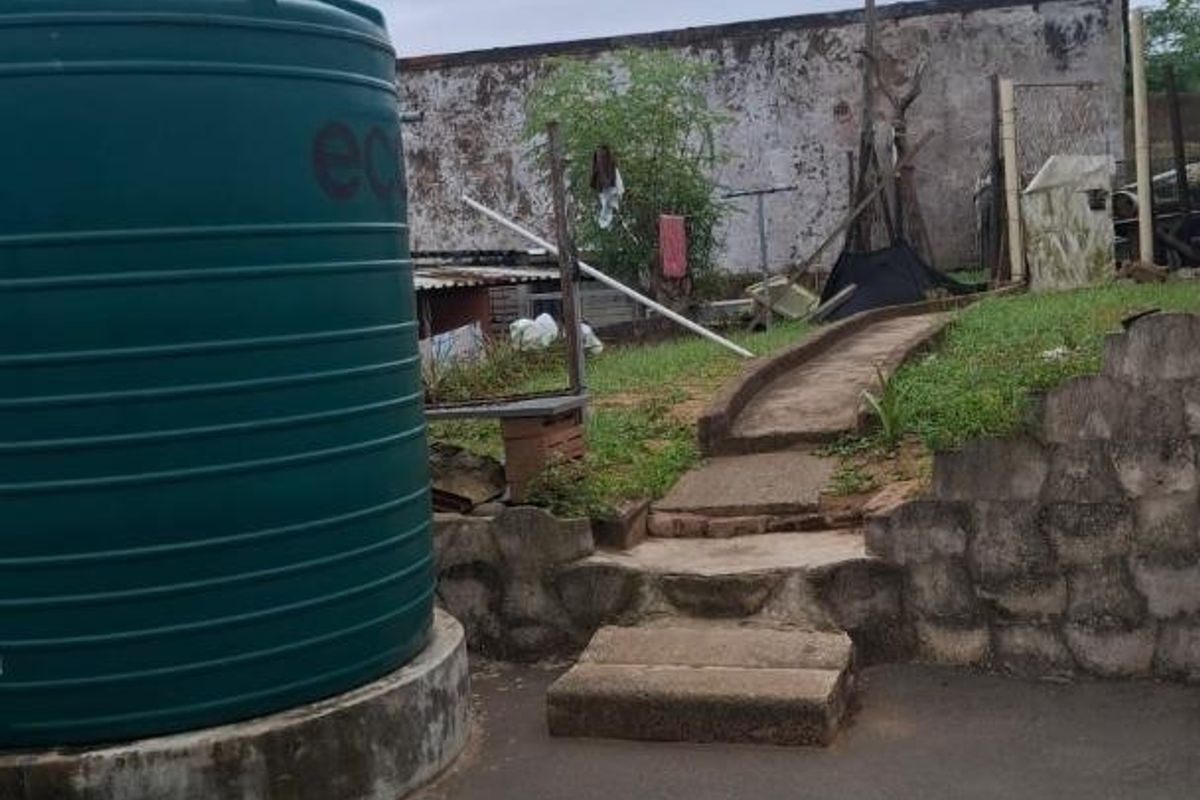 Rear exterior area featuring a large water storage tank, concrete steps leading to a raised garden section, and a weathered boundary wall.