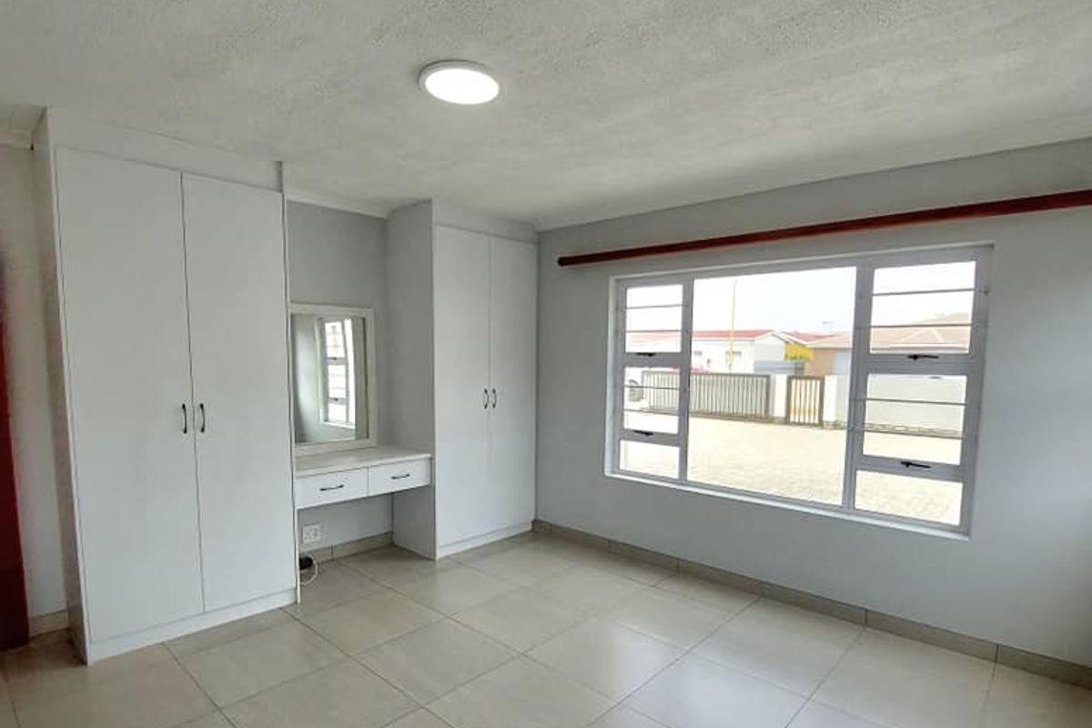 A bright bedroom featuring neutral floor tiles, built-in cupboards with an integrated vanity, and large windows providing natural light.