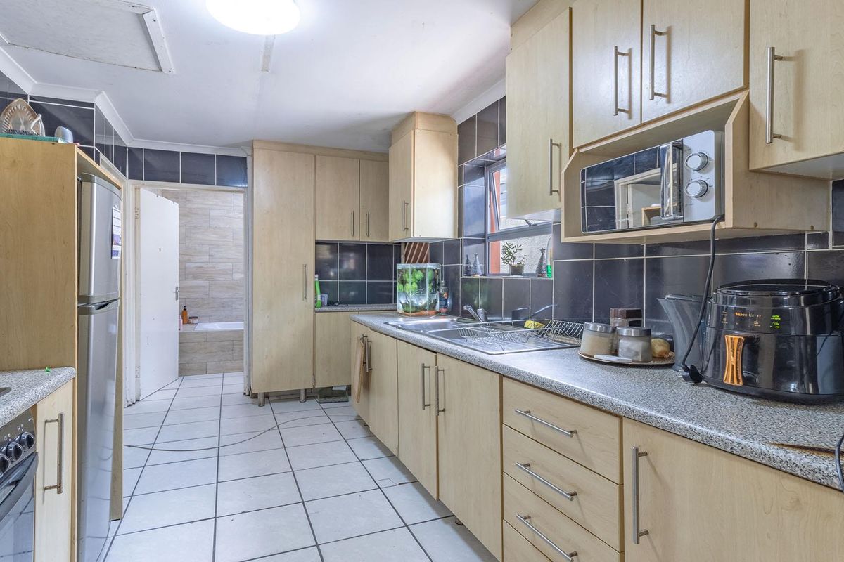 A functional kitchen featuring light wood-toned cabinetry, dark tiled splashbacks, stone-look countertops, and tiled flooring, with an adjacent view into the bathroom.
