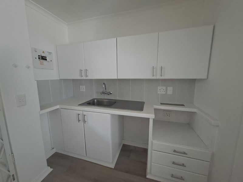 A compact, functional kitchen area featuring white built-in cabinetry, a stainless steel sink, and a tiled backsplash with neutral laminate flooring.
