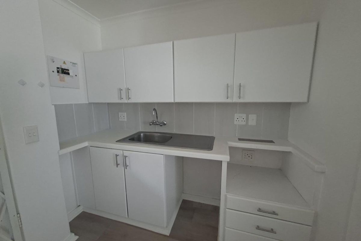 A compact, functional kitchen area featuring white built-in cabinetry, a stainless steel sink, and a tiled backsplash with neutral laminate flooring.