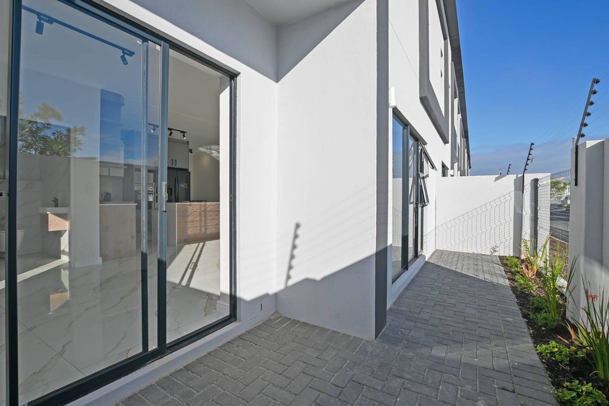 A modern rear exterior view of the townhouse featuring a paved patio area, sliding glass doors, and secure boundary wall with electric fencing.