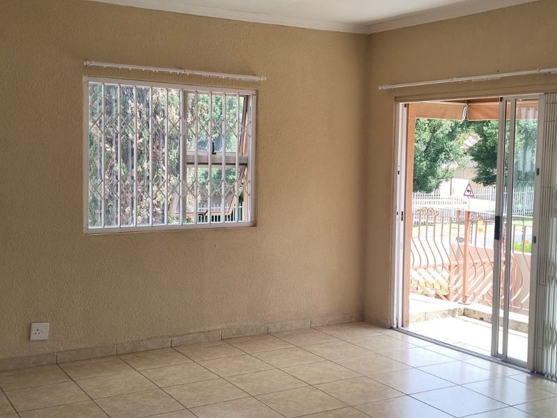A spacious, empty living room featuring neutral tiled flooring, large windows with security bars, and a sliding door leading to an outdoor area.