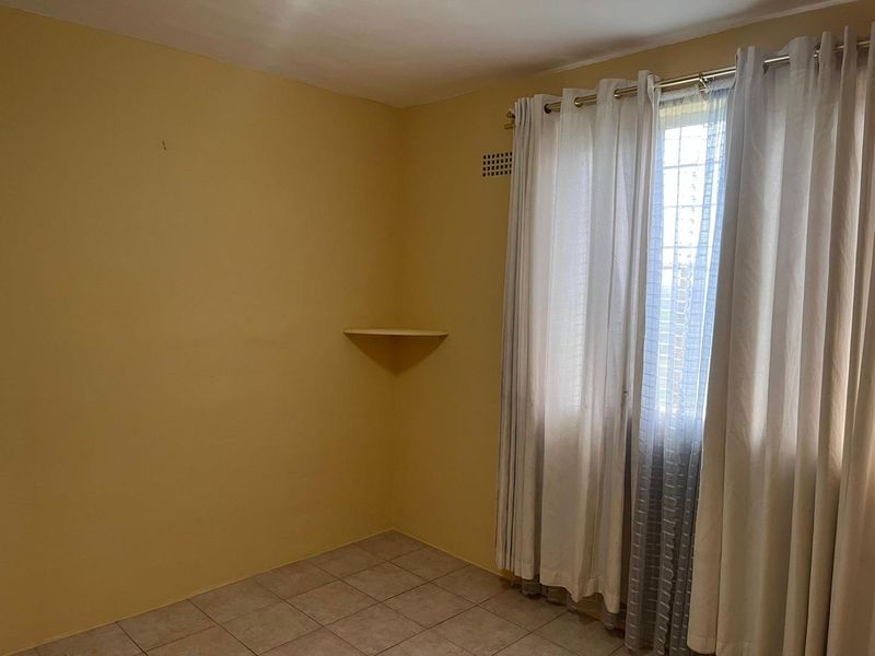 A vacant bedroom featuring tiled flooring, a built-in corner shelf, and a window fitted with security bars.