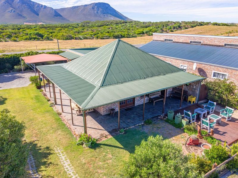 Aerial view of a brick house featuring a large wrap-around covered patio, corrugated metal roof, timber decking, and expansive surrounding garden and landscape.