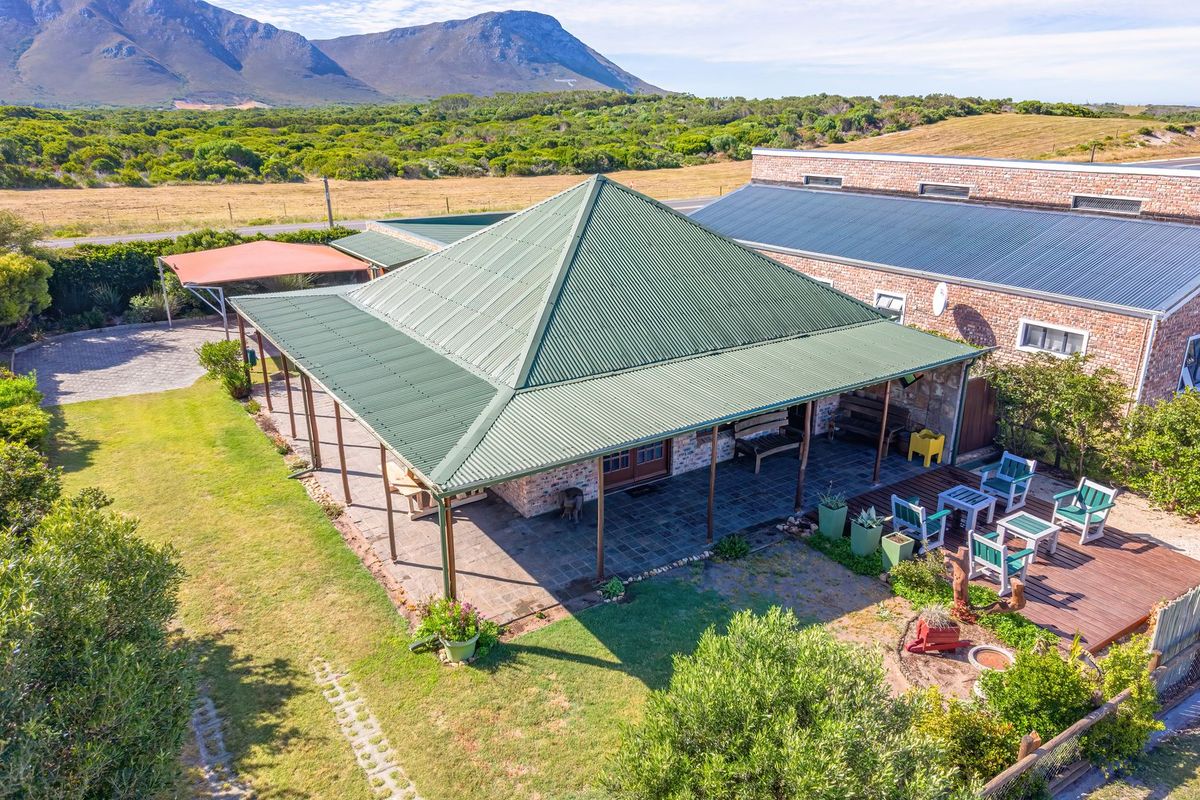 Aerial view of a brick house featuring a large wrap-around covered patio, corrugated metal roof, timber decking, and expansive surrounding garden and landscape.