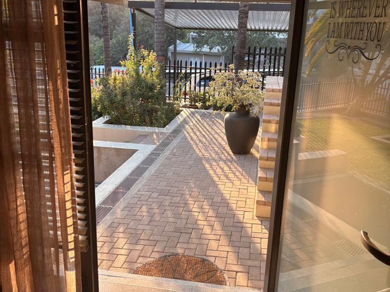 A view from an interior space onto a paved outdoor patio area featuring a covered pergola, retaining walls, and secure perimeter fencing.