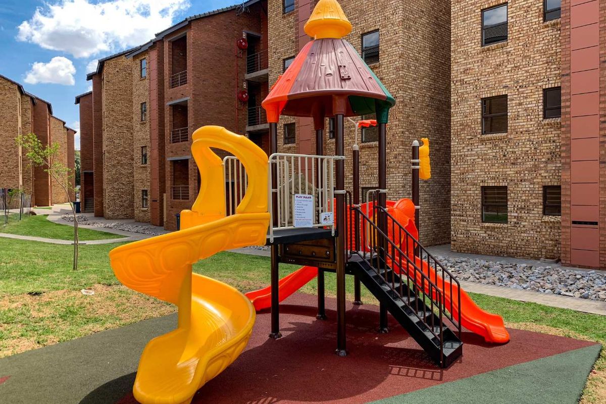 A well-maintained communal playground area situated between face brick apartment blocks, featuring modern play equipment and landscaped grounds.