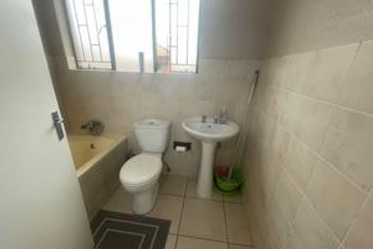 A functional bathroom featuring a bathtub, pedestal basin, toilet, and neutral wall and floor tiling with security bars on the window.