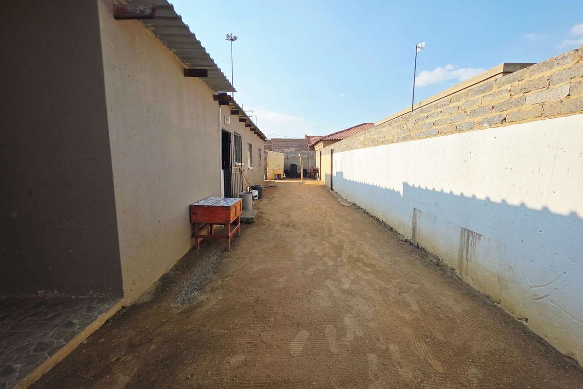 A side exterior view of the property featuring a long, unpaved pathway enclosed by a high boundary wall and the side of the house.