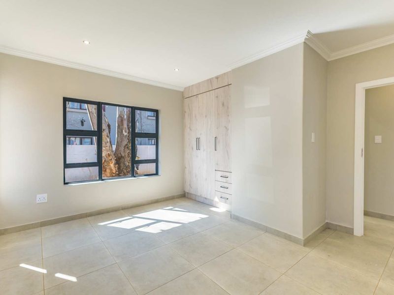 A bright, empty bedroom featuring neutral tiled flooring, modern built-in cupboards, and large aluminium-framed windows providing natural light.