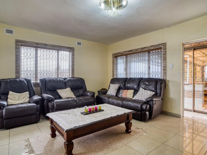 A spacious living room featuring tiled flooring, a decorative crystal chandelier, and large windows fitted with security bars, offering access to an adjacent patio area.