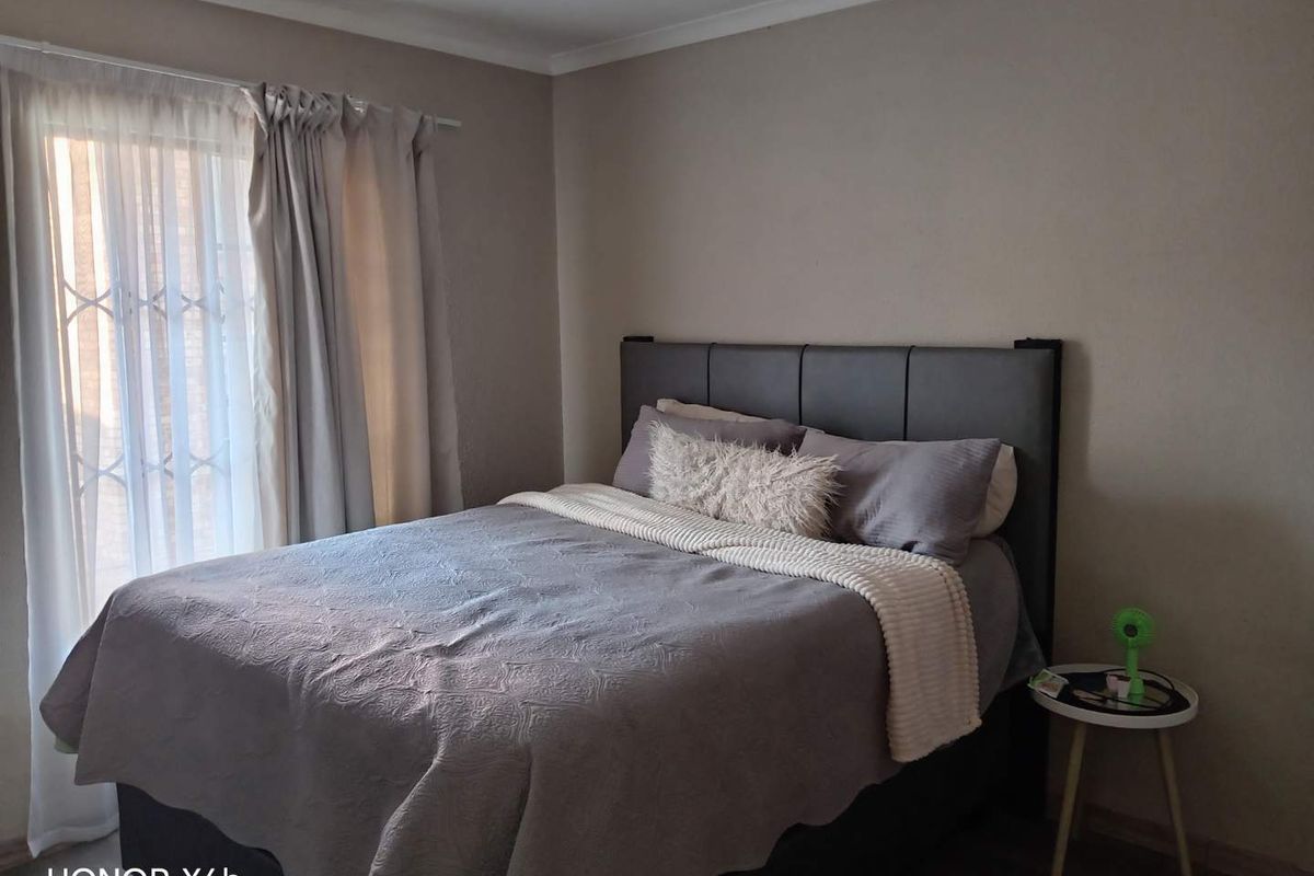 A neutrally painted bedroom featuring a large window with a security gate and curtain rod, providing natural light and a functional living space.