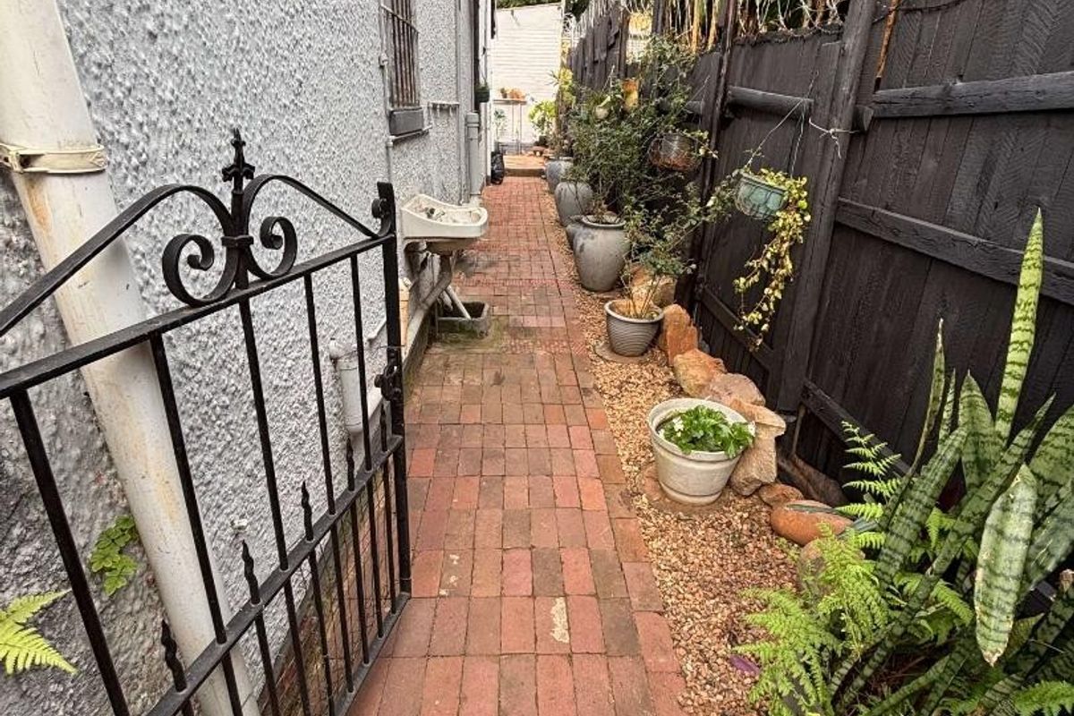 A narrow side passage featuring brick paving, a wrought iron gate, and a wall-mounted utility basin along the exterior wall.