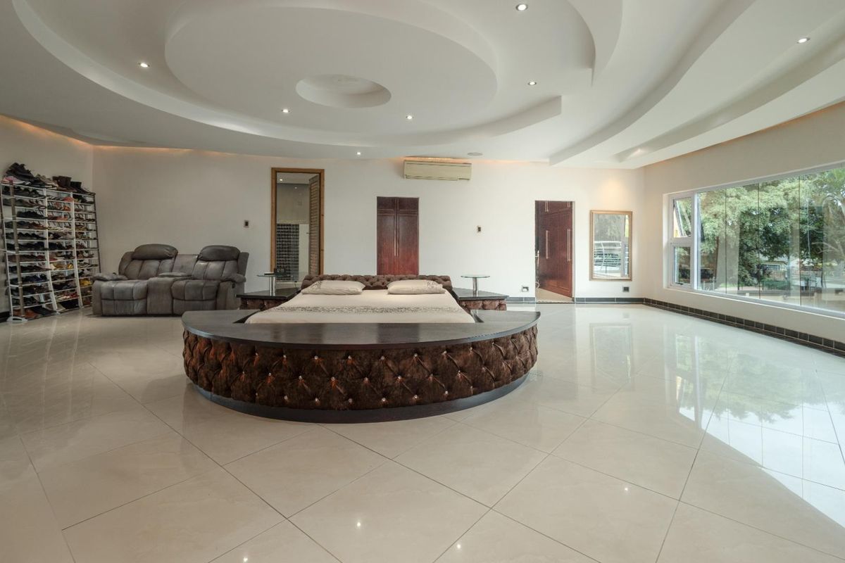 Spacious master bedroom featuring a unique circular bed platform, intricate coffered ceiling with recessed lighting, and large windows overlooking the garden.