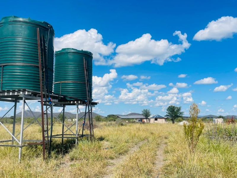 A rural farm property featuring elevated green water storage tanks on a metal stand, set within an open landscape with distant structures.