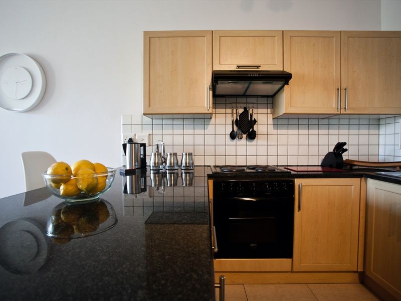 A functional kitchen featuring light-wood cabinetry, black granite countertops, a tiled backsplash, and integrated black appliances.