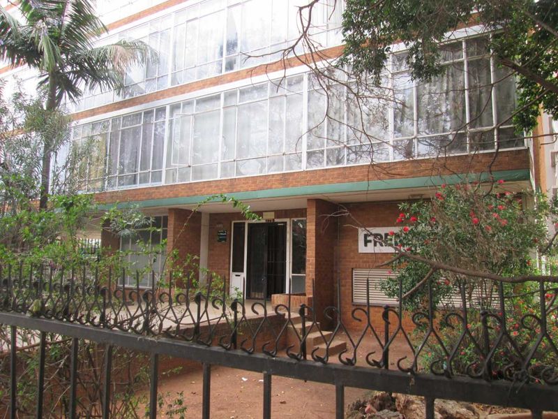 Front elevation of a multi-story apartment building featuring brick facade, extensive glass window panels, and a secure wrought iron perimeter fence.