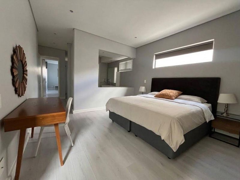 A modern bedroom featuring light-toned laminate flooring, recessed ceiling lighting, and a unique internal window providing a view into the adjacent bathroom.