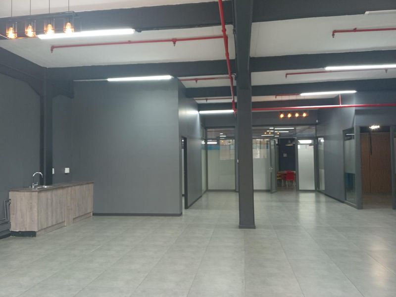Spacious open-plan office area featuring modern grey finishes, exposed industrial-style ceiling elements, tiled flooring, and a convenient kitchenette corner.