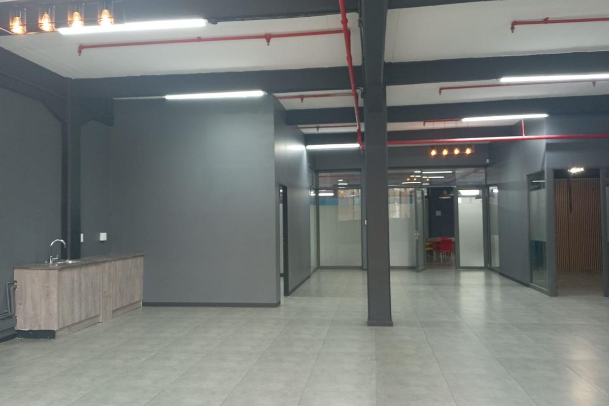 Spacious open-plan office area featuring modern grey finishes, exposed industrial-style ceiling elements, tiled flooring, and a convenient kitchenette corner.