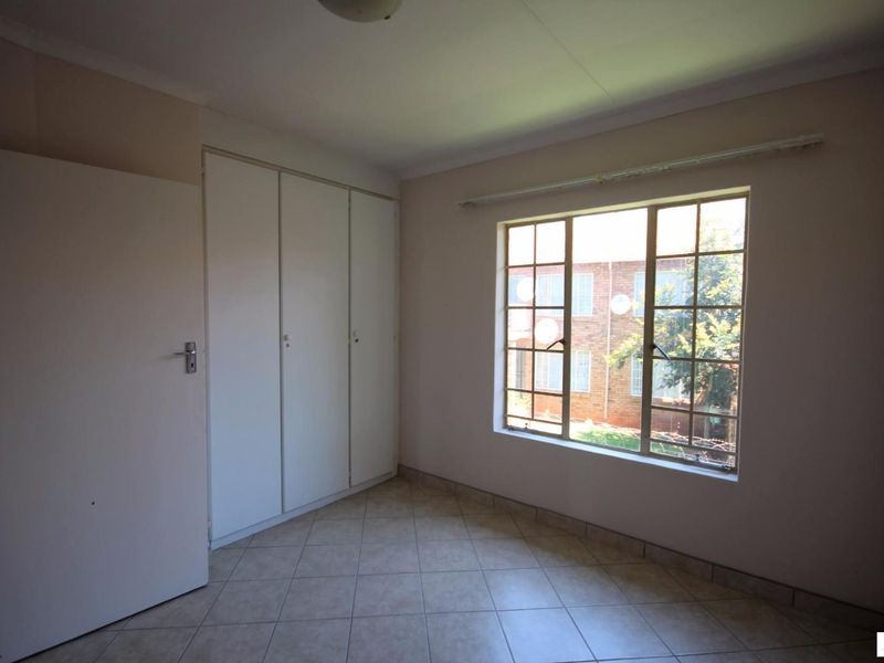 A bright, empty bedroom featuring tiled flooring, built-in cupboards, and a window with security bars.