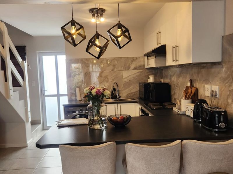 Modern open-plan kitchen featuring a breakfast bar, stone-look backsplash, built-in cabinetry, and stylish pendant lighting, seamlessly connected to the living area and staircase.