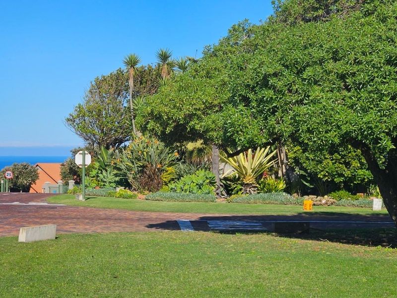 A view of the street approach featuring a paved driveway, well-maintained landscaped garden, and mature trees with a distant ocean view.