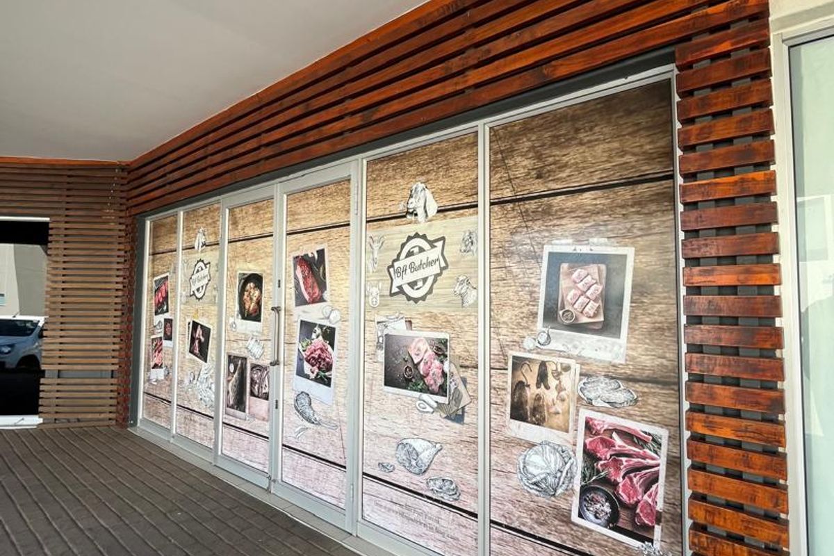 Front facade of a commercial butcher shop featuring timber slat detailing, paved walkway, and large glass storefront with branded window graphics.
