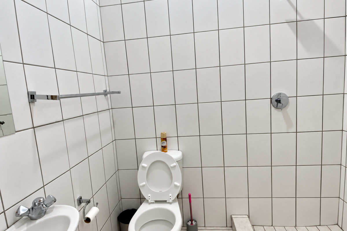A functional bathroom featuring white wall tiles, a standard toilet, a wall-mounted shower head, and a compact washbasin with chrome fixtures.