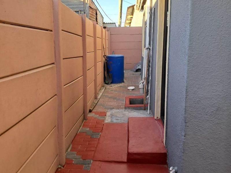 A narrow side passage featuring a precast concrete boundary wall, paved walkway, and a small protective awning over the entrance.