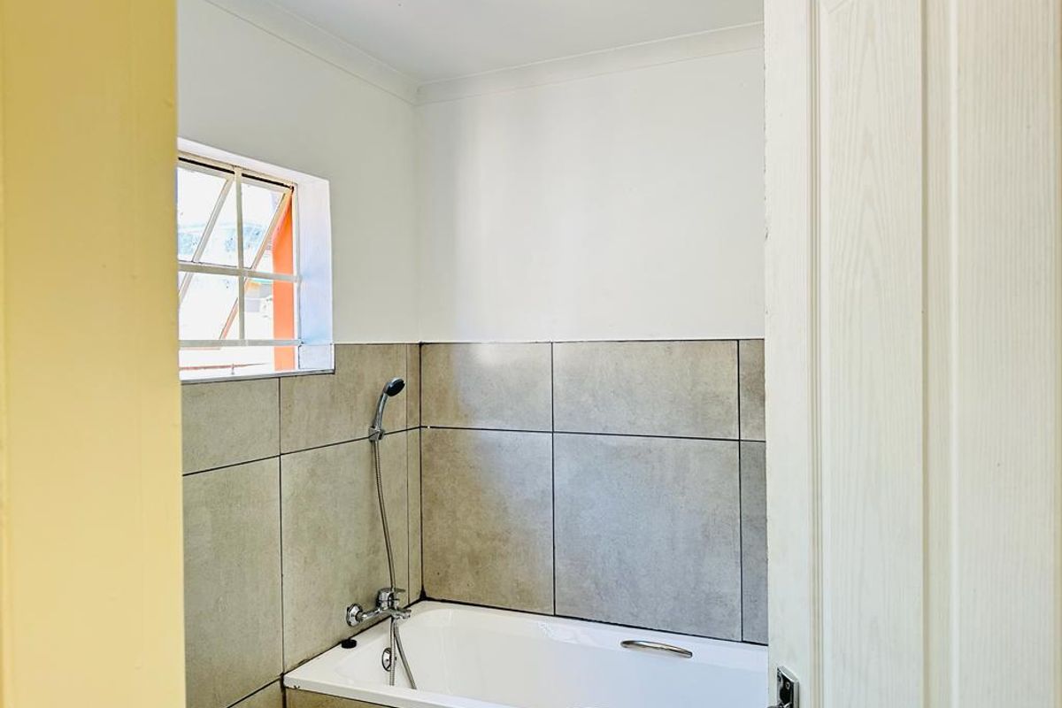 A functional bathroom featuring a built-in bathtub with a handheld shower mixer, complemented by neutral wall tiling and a small window for natural light.