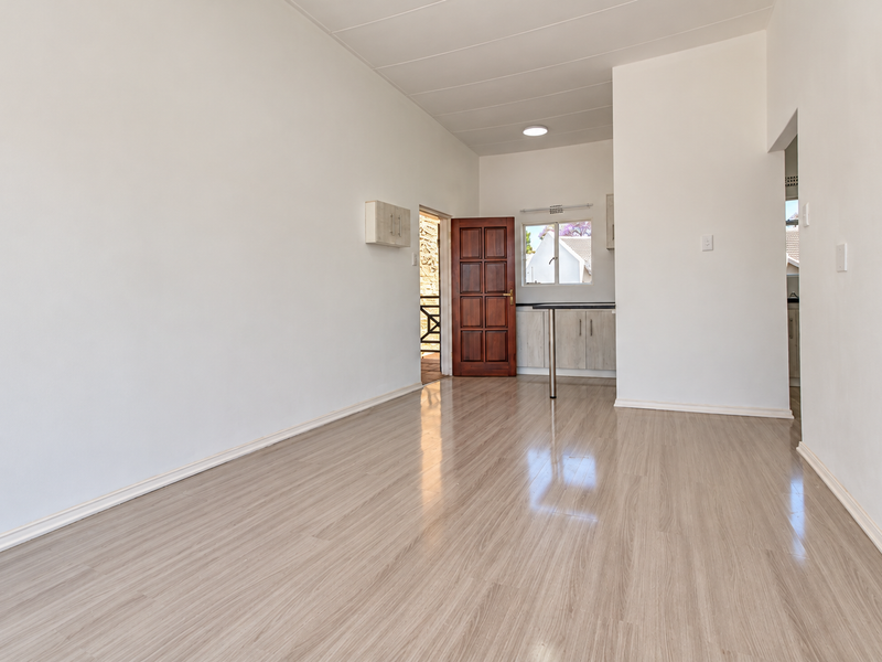 An empty open-plan living and kitchen area featuring light-toned laminate flooring, neutral walls, and built-in kitchen cabinetry with a breakfast counter.