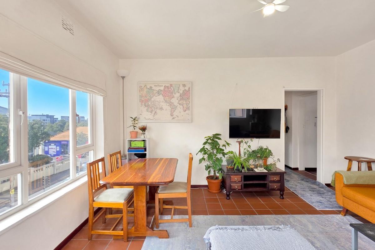 A bright living room featuring terracotta floor tiles, a ceiling fan, and large windows providing natural light and an urban view.